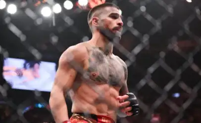 Muscular male mixed martial arts fighter with tattoos, wearing red shorts and gloves, standing inside a cage.