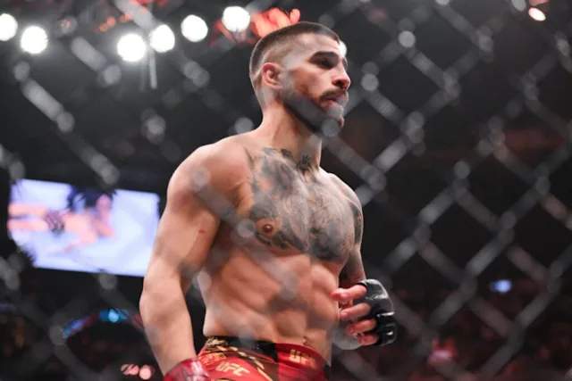Muscular male mixed martial arts fighter with tattoos, wearing red shorts and gloves, standing inside a cage.