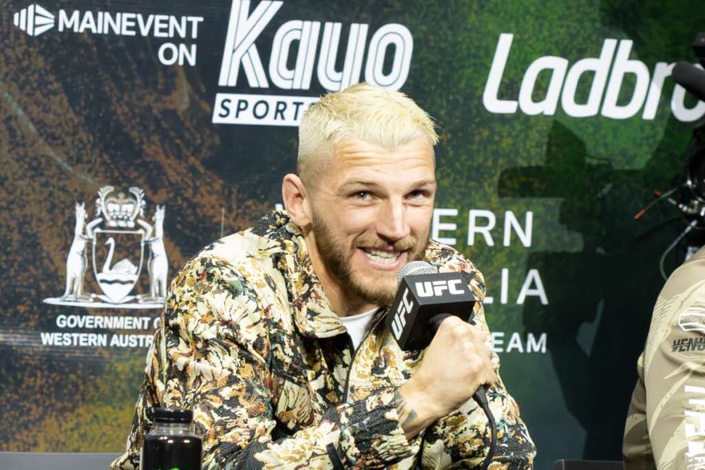 Man with bleached blond hair in a floral shirt speaks into a UFC microphone at a press conference backdrop.
