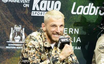 Man with bleached blond hair in a floral shirt speaks into a UFC microphone at a press conference backdrop.