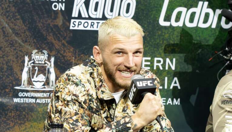 Man with bleached blond hair in a floral shirt speaks into a UFC microphone at a press conference backdrop.