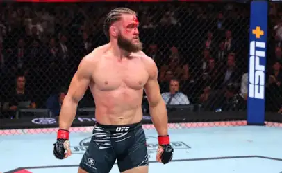 Male mixed martial artist stands in the UFC octagon with blood on his forehead, wearing black UFC shorts and red gloves, looking to the left.