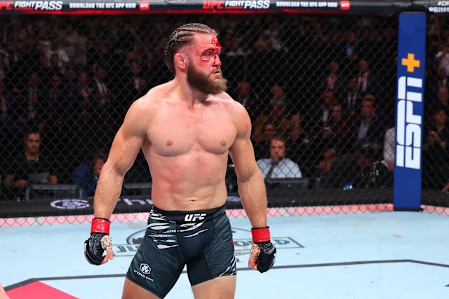 Male mixed martial artist stands in the UFC octagon with blood on his forehead, wearing black UFC shorts and red gloves, looking to the left.