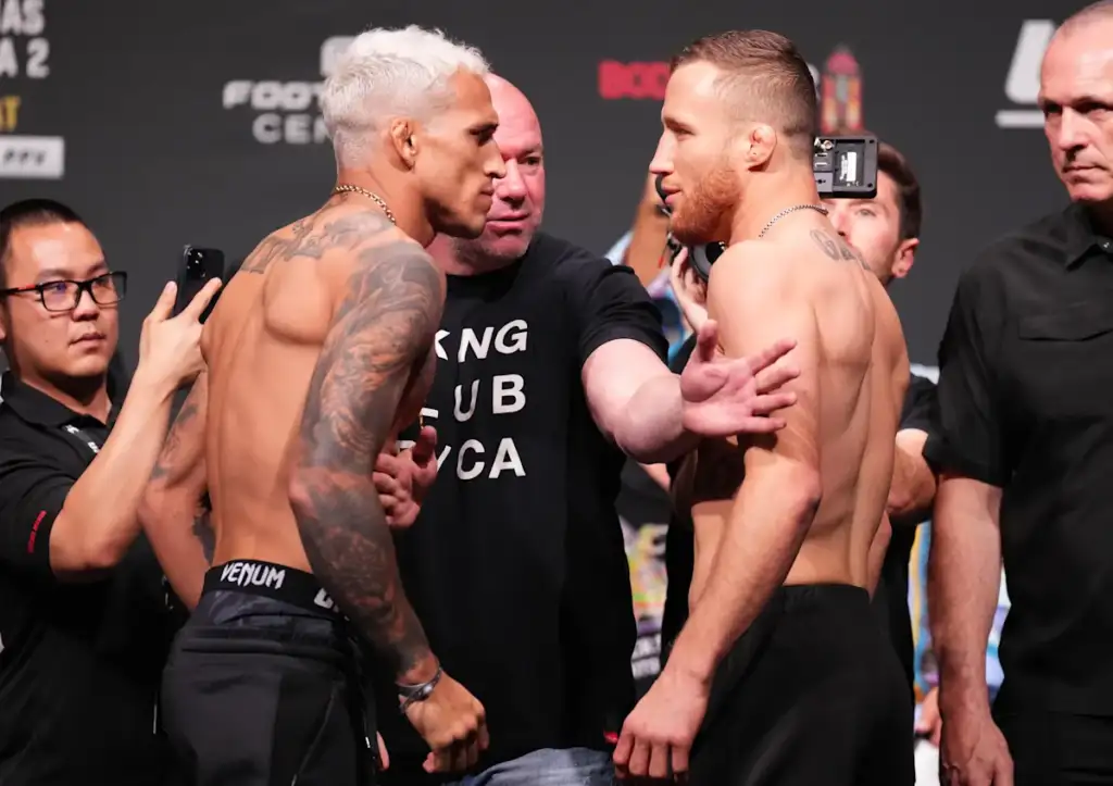 Two shirtless fighters face off on a stage during a weigh-in as photographers document the moment Bring between them a man in a black shirt holds them apart.