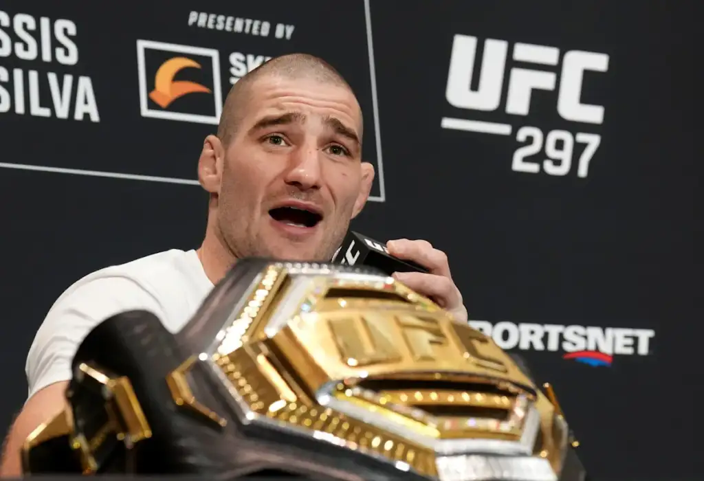 Male MMA fighter speaks into a microphone at a post-fight press conference, holding a shiny gold UFC championship belt in front of a black backdrop with UFC logos and 'UFC 297' text from SportsNet signage