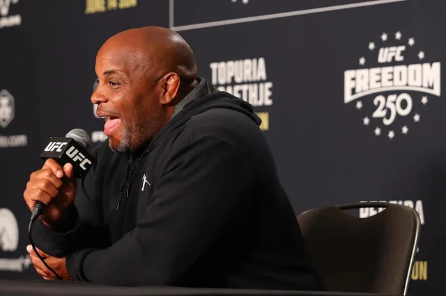 Bald man in a black hoodie speaks into a UFC microphone at a press conference backdrop.