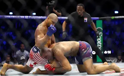Two male mixed martial artists grapple on the mat inside a fenced cage; one in red‑white striped shorts with red gloves pins the other in blue striped shorts. A referee watches from behind.