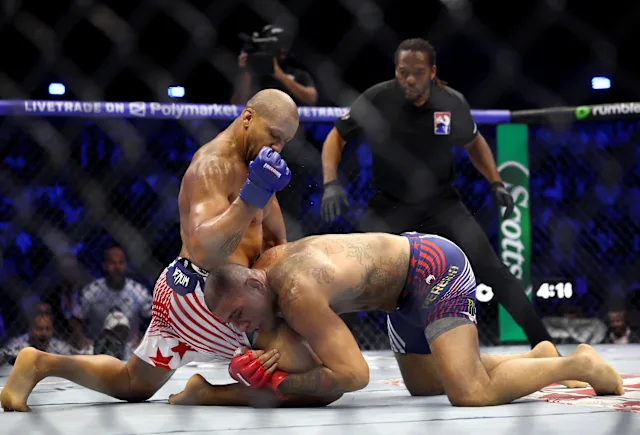 Two male mixed martial artists grapple on the mat inside a fenced cage; one in red‑white striped shorts with red gloves pins the other in blue striped shorts. A referee watches from behind.