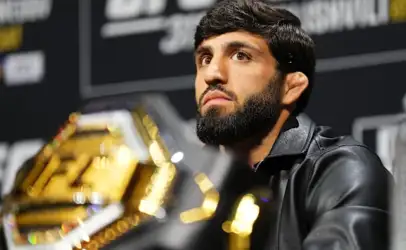 Male mixed martial artist with a beard at a press conference, UFC belt partly visible in the foreground.