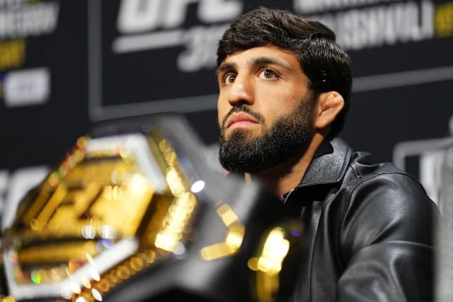 Male mixed martial artist with a beard at a press conference, UFC belt partly visible in the foreground.