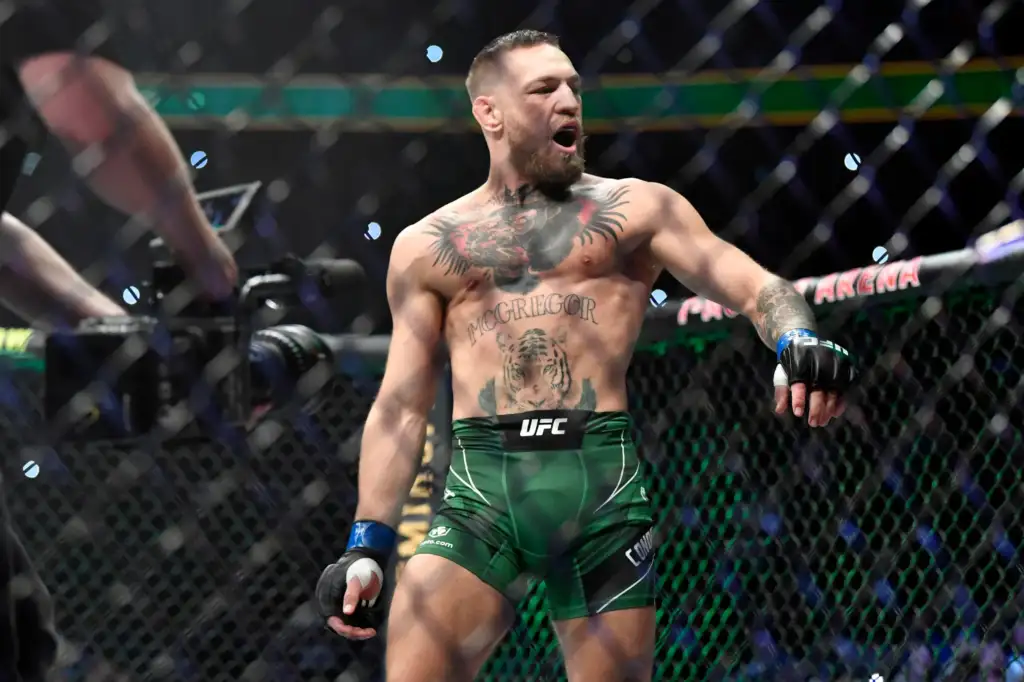 Male UFC fighter with tattoos celebrating inside the octagon, shirtless, wearing green shorts, fists clenched in triumph.