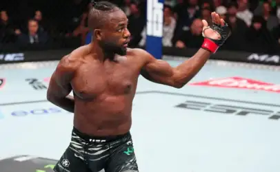 Male mixed martial artist inside the UFC cage raises his right arm in victory after a match, wearing black fight shorts and red wrist wraps.