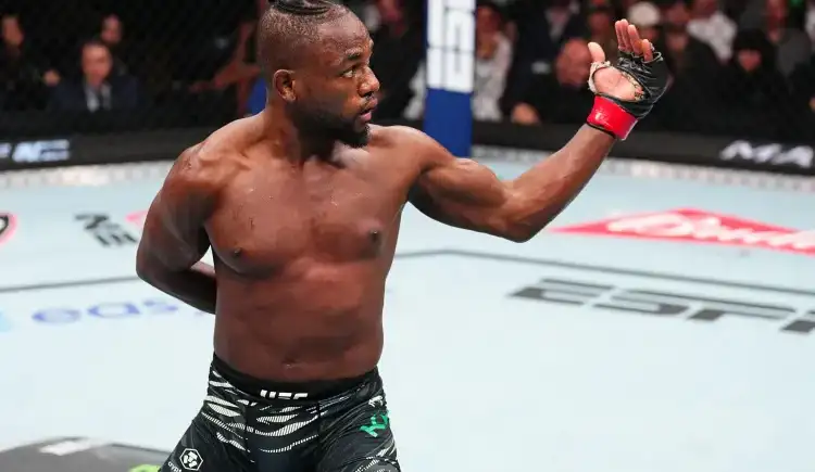Male mixed martial artist inside the UFC cage raises his right arm in victory after a match, wearing black fight shorts and red wrist wraps.