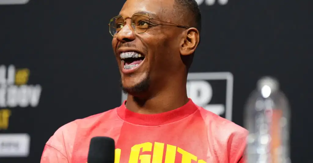 gettyimages-2194304438 - AfroCombat Man wearing glasses and a pink shirt, speaking into a microphone with a big smile during a public event.