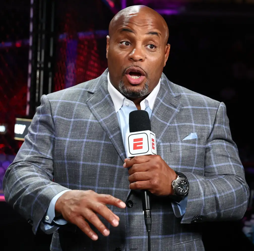 gettyimages-2230500946 - AfroCombat Man in a gray plaid suit speaks into a handheld microphone with a surprised facial expression on a TV set.
