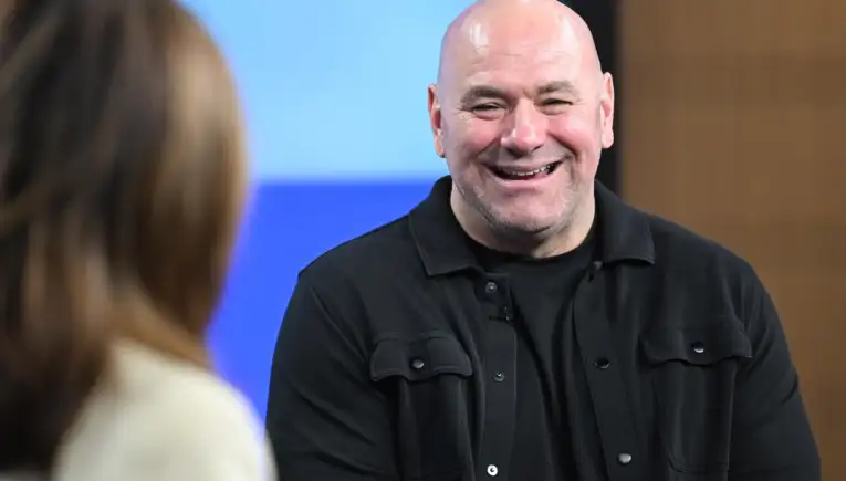 Smiling bald man in a black shirt talks to someone off-camera at an indoor event.