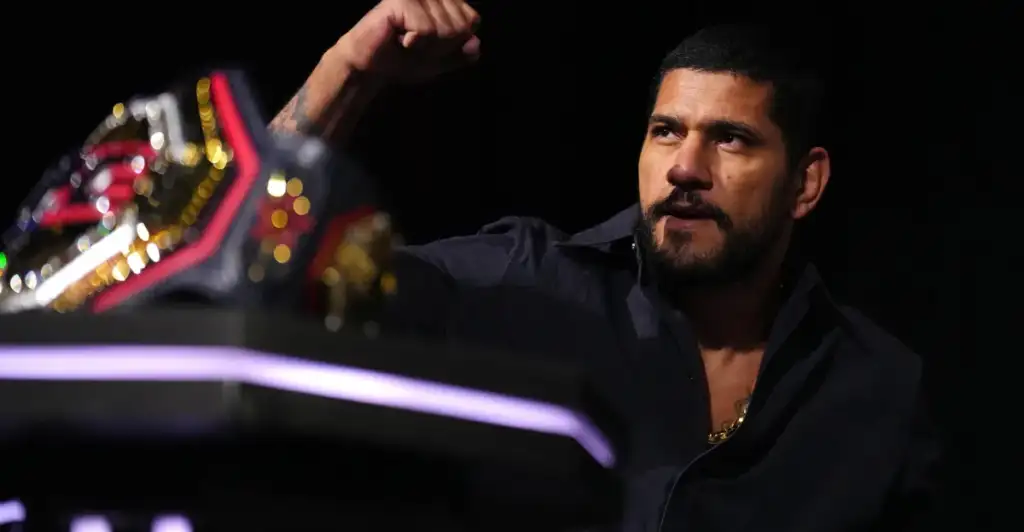 Male wrestler with a raised fist, gazing off to the right, championship belt visible in the foreground by stage lights.