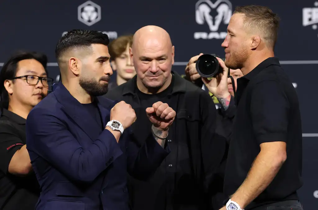 Two men in a stare-down pose with fists raised, as a bald man watches and photographers capture the moment in a dark backdrop.