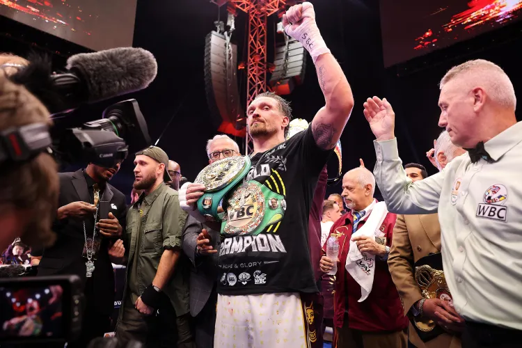 Boxer with two championship belts raises his arm in victory as cameras and teammates celebrate ringside.