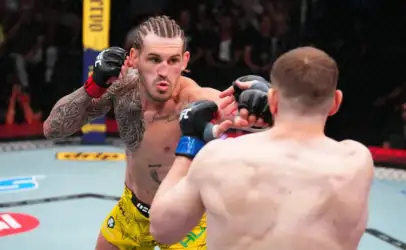 MMA fighter with braided hair and tattoos throws a punch toward his opponent in the cage, wearing yellow trunks and black gloves.