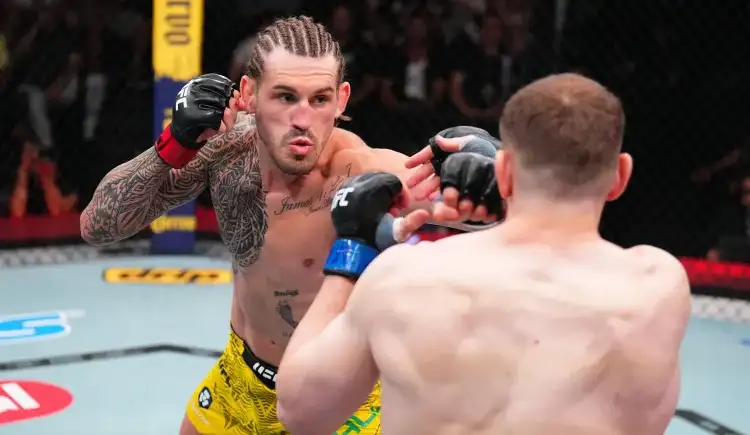 MMA fighter with braided hair and tattoos throws a punch toward his opponent in the cage, wearing yellow trunks and black gloves.