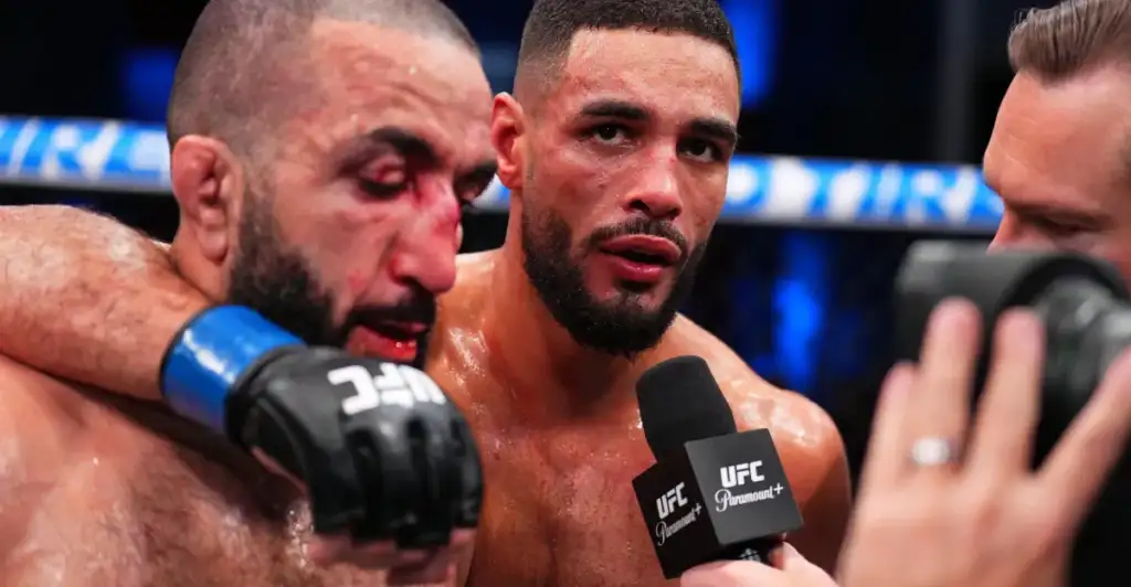 Two UFC fighters speak to a reporter in the ring; one fighter with a bloody face is being interviewed while another holds him close, wearing gloves and taped wrists.