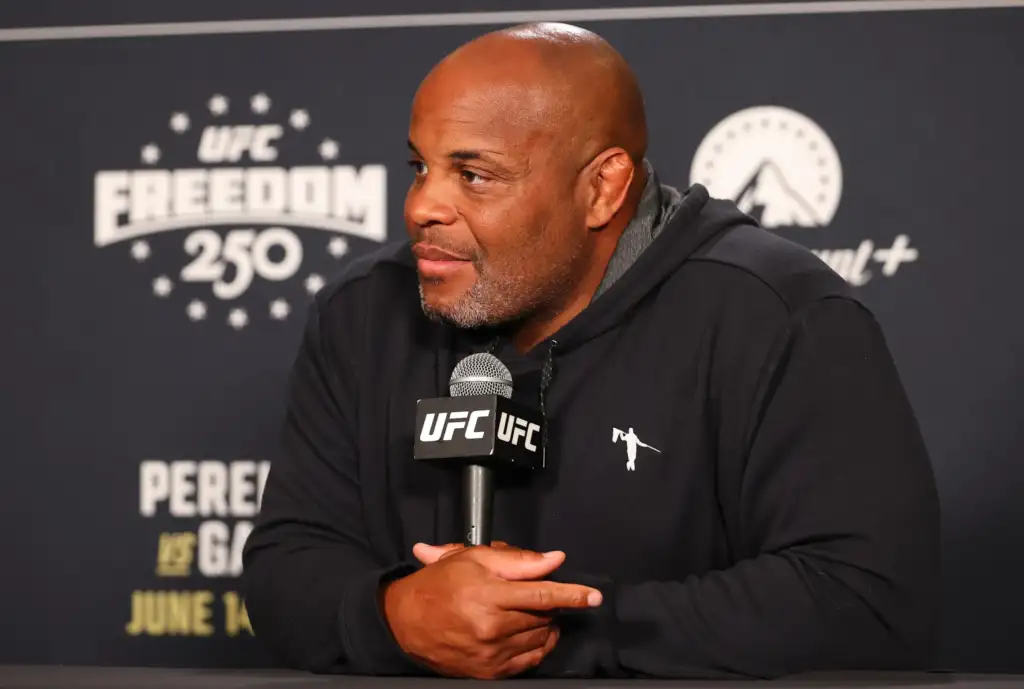 Man in a black hoodie at a UFC press conference, hands folded around a UFC microphone, looking to the side.