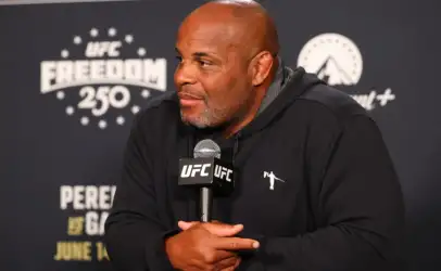 Man in a black hoodie at a UFC press conference, hands folded around a UFC microphone, looking to the side.