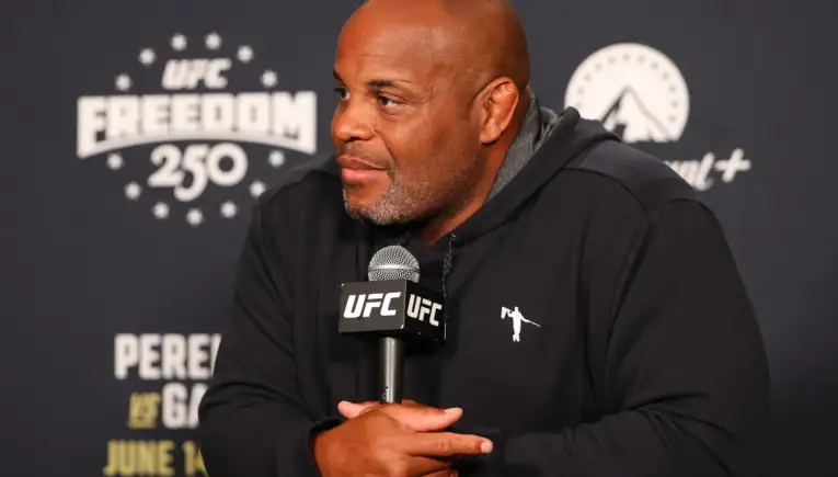 Man in a black hoodie at a UFC press conference, hands folded around a UFC microphone, looking to the side.