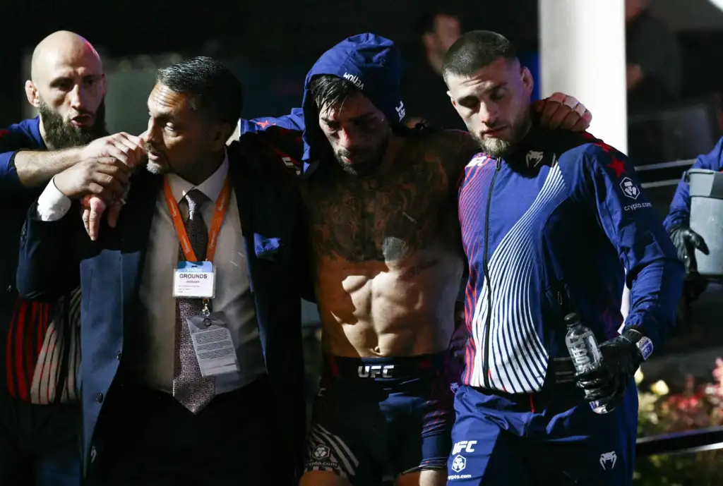 Group of men with UFC gear standing arm-in-arm backstage after a fight; one shirtless fighter with tattoos supported by teammates in blue jackets and lanyards.