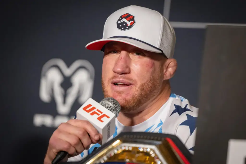 Male mixed martial artist wearing a white cap with a logo speaks into a UFC microphone at a press event, championship belt visible in foreground