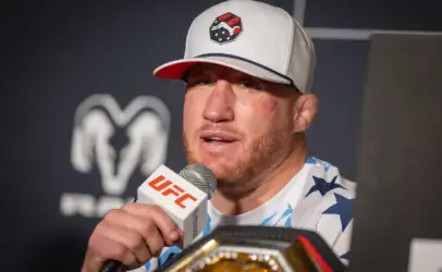 Male mixed martial artist wearing a white cap with a logo speaks into a UFC microphone at a press event, championship belt visible in foreground