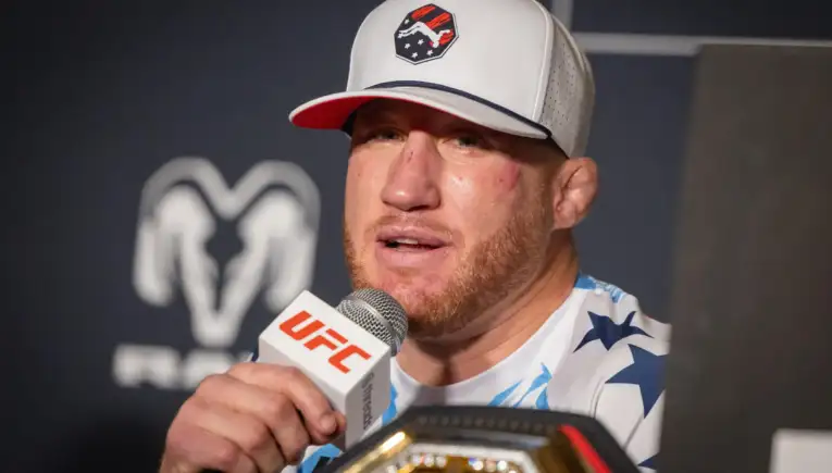 Male mixed martial artist wearing a white cap with a logo speaks into a UFC microphone at a press event, championship belt visible in foreground