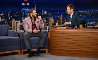 Talk show set with host behind a curved wooden desk presenting a plaque to a smiling guest in a pink checked blazer seated on a blue sofa, NYC skyline backdrop behind them.