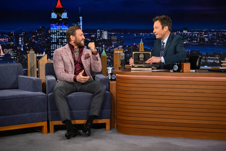 gettyimages-2281357737 - AfroCombat Talk show set with host behind a curved wooden desk presenting a plaque to a smiling guest in a pink checked blazer seated on a blue sofa, NYC skyline backdrop behind them.