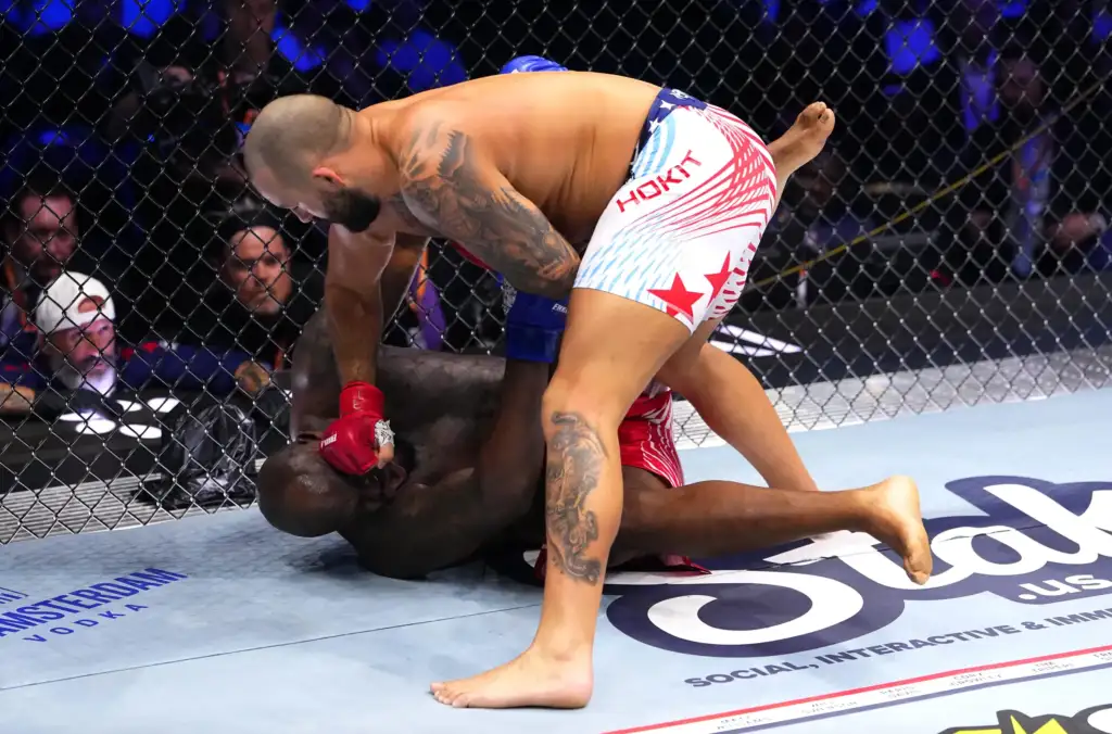 gettyimages-2281622060 - AfroCombat Two mixed martial artists grapple on the mat inside a cage; one fighter in red gloves pins the other, with spectators visible in the background.