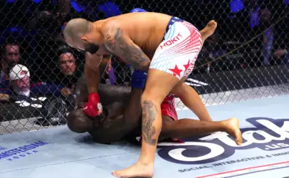 Two mixed martial artists grapple on the mat inside a cage; one fighter in red gloves pins the other, with spectators visible in the background.