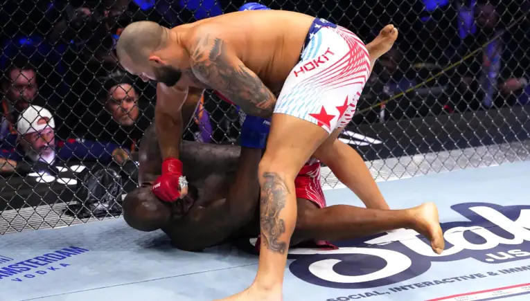 Two mixed martial artists grapple on the mat inside a cage; one fighter in red gloves pins the other, with spectators visible in the background.