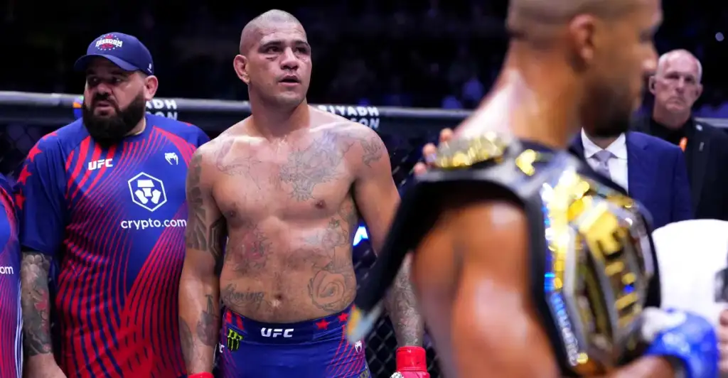 Tattooed UFC fighter with red gloves stands shirtless in the octagon, with teammates nearby and a belt-wearing champion blurred in the foreground.