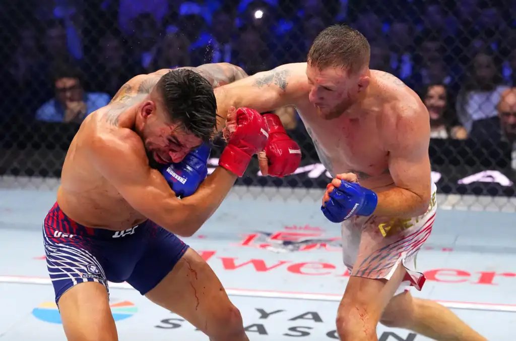 Two mixed martial artists exchange punches and clinch inside a cage during a bout, sweat and blood visible on their bodies.