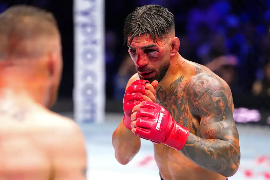 A bruised, tattooed fighter in red gloves inside a ring, hands up in a defensive stance.