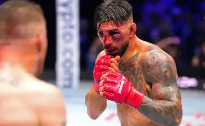 A bruised, tattooed fighter in red gloves inside a ring, hands up in a defensive stance.