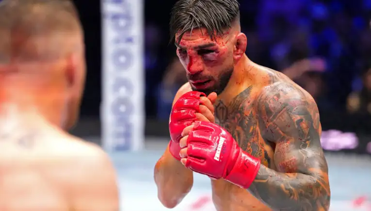 A bruised, tattooed fighter in red gloves inside a ring, hands up in a defensive stance.