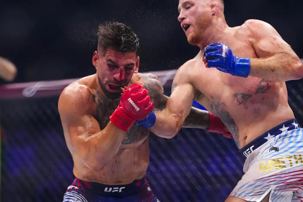 Two shirtless mixed martial artists exchange punches in a cage; fighter with red gloves lands a blow on a bleeding opponent with blue gloves nearby.