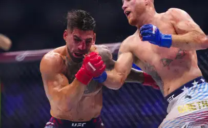 Two shirtless mixed martial artists exchange punches in a cage; fighter with red gloves lands a blow on a bleeding opponent with blue gloves nearby.