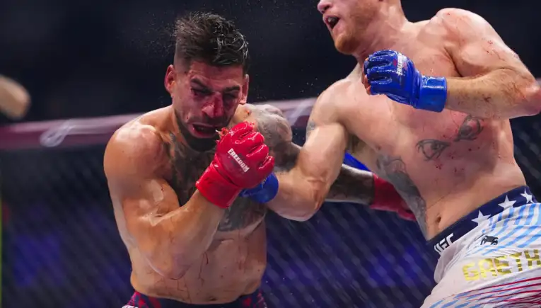 Two shirtless mixed martial artists exchange punches in a cage; fighter with red gloves lands a blow on a bleeding opponent with blue gloves nearby.