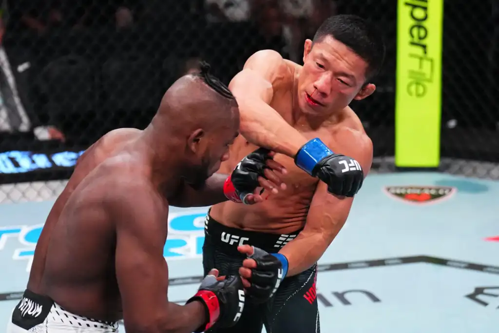 Two shirtless UFC fighters clash inside the octagon as one punches the other in the chest, both wearing gloves and intense expressions.