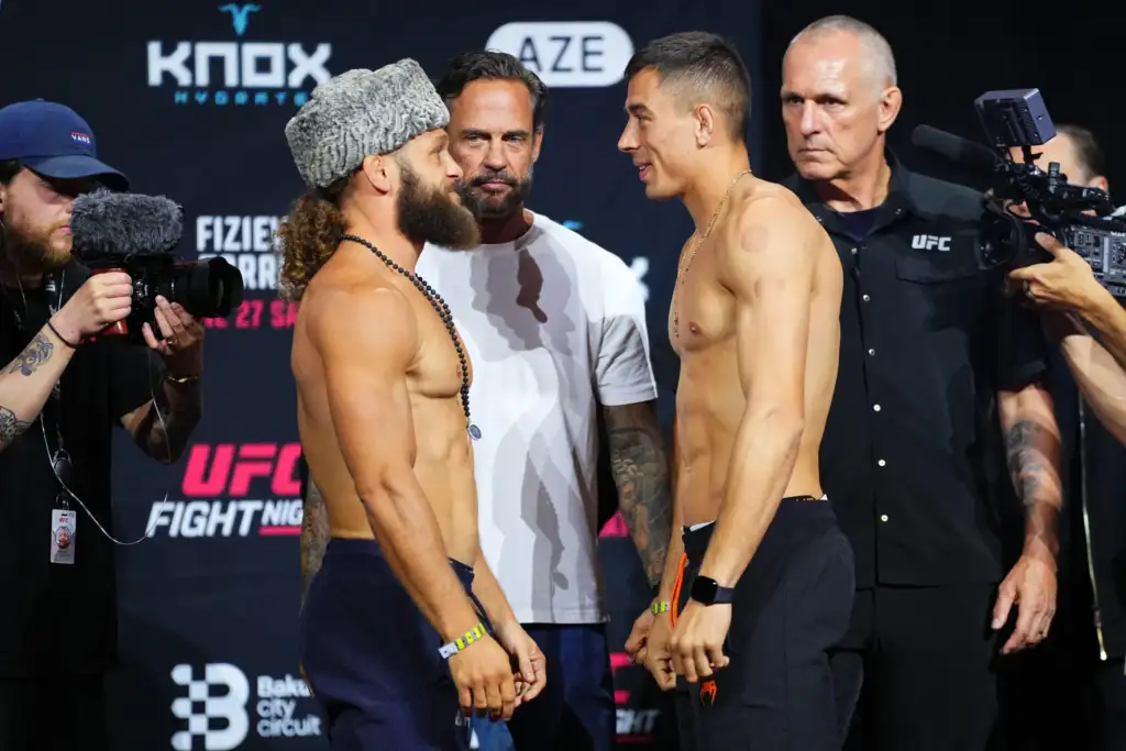 Two shirtless fighters face off in a crowded weigh-in stage, eyes locked, with cameras and crew surrounding them and a UFC backdrop behind them.