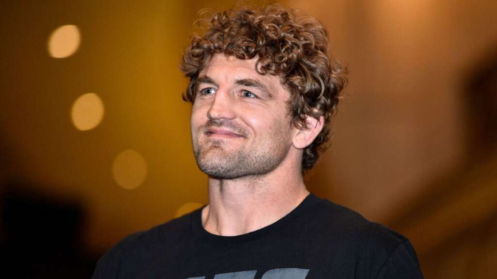 Close-up of a smiling man with curly brown hair wearing a black T-shirt against a warm bokeh background.