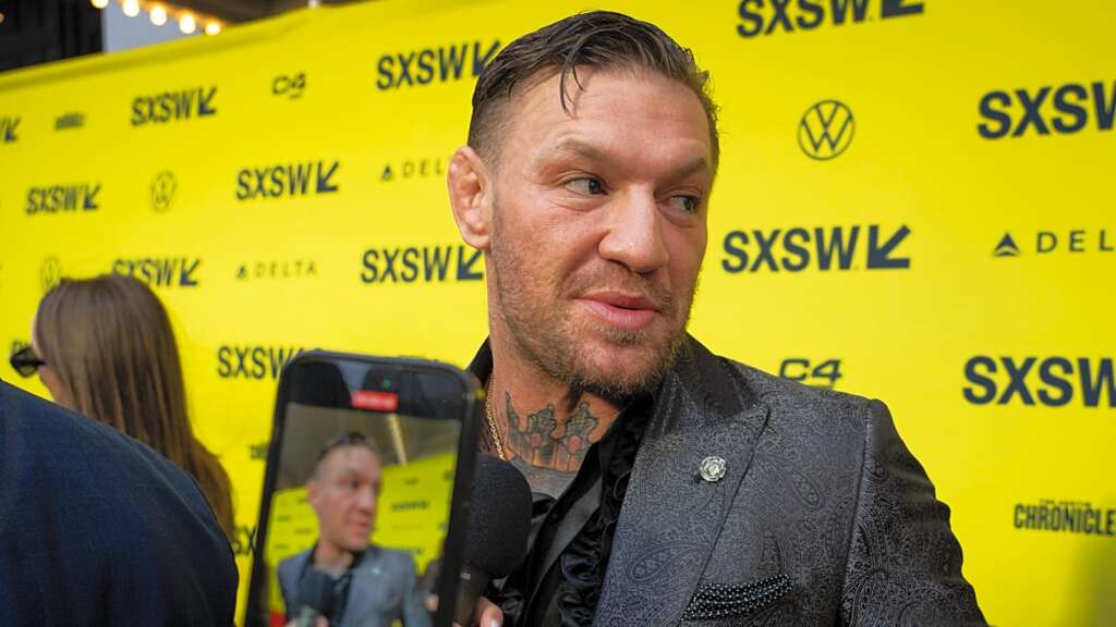 march-8-2024-austin-tx-conor-mcgregor-speaks - AfroCombat Man in a patterned blazer being interviewed at SXSW against a bright yellow backdrop filled with SXSW logos.